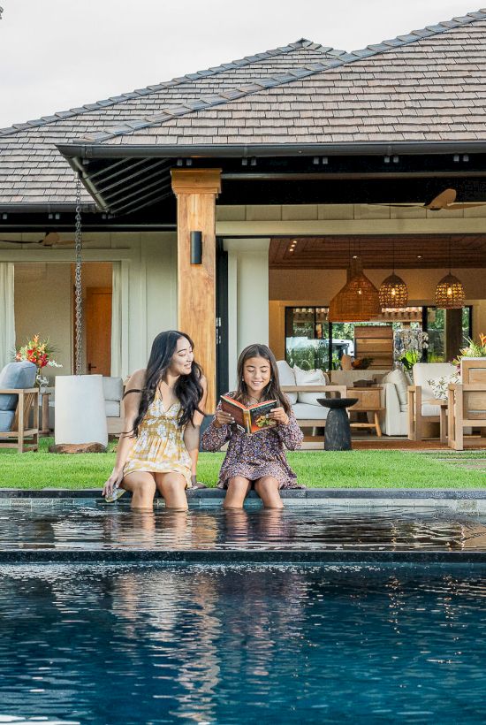 A man greets a family on a patio by a pool where two kids sit and read while water ripples.