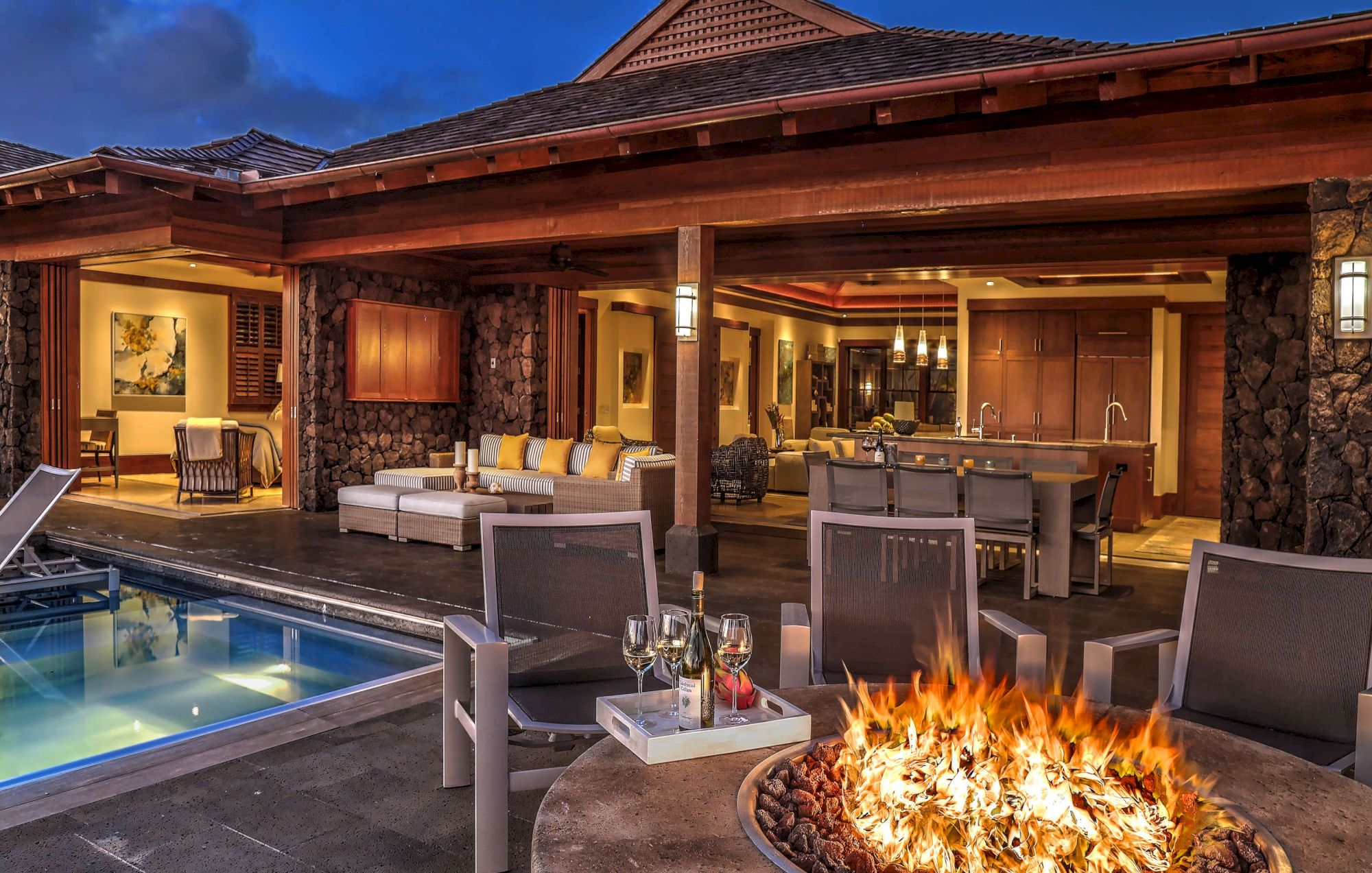 Luxury outdoor patio with a swimming pool, stone fire pit flames, lounge seating, and a built-in bar/kitchen under a wooden roof.