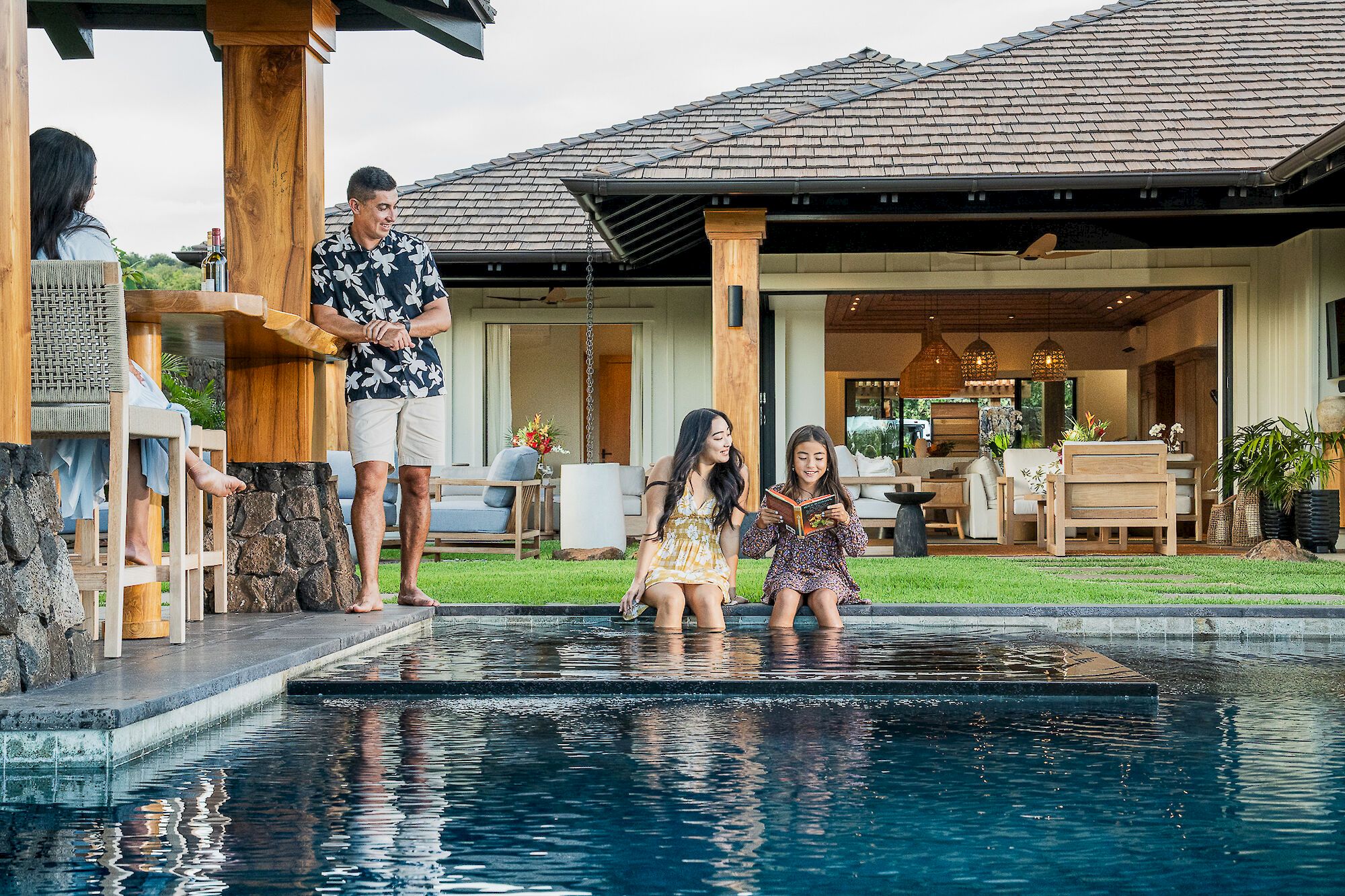 A man stands by a resort pool while two girls sit and read by the patio, surrounded by wooden pillars, furniture, and greenery.