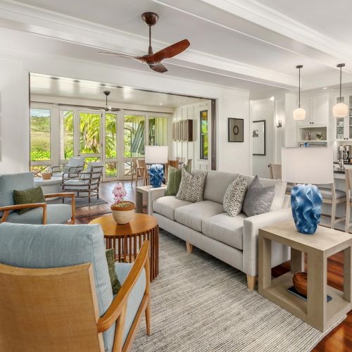 Bright open-plan living room with sofa, coffee tables, and a white kitchen with island and stools, plus large windows.