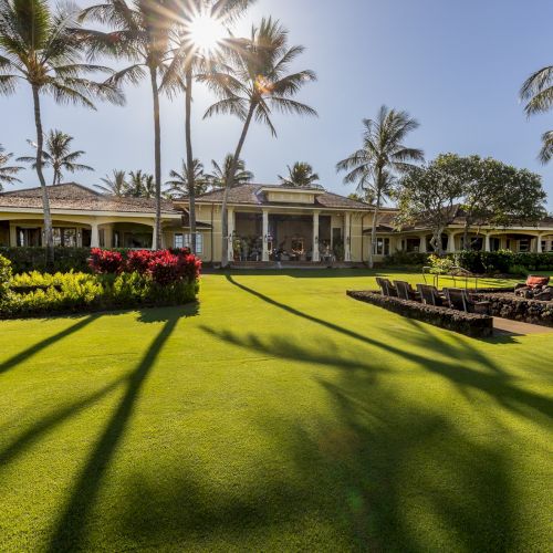 A sunny resort courtyard with a manicured green lawn, palm trees, colorful flowerbeds, and a long beige building with seating in front.