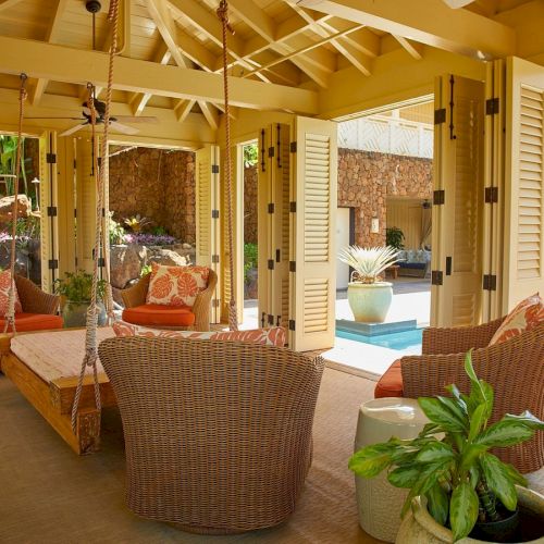 Sunlit covered patio with wicker swings and seating, tropical plants, and a glimpse of a swimming pool beyond open doors.