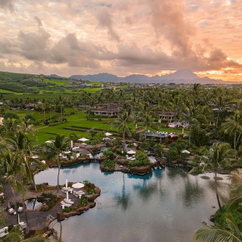 A tropical resort landscape with lush palm trees, a winding lagoon, and green grounds under a dramatic sunset sky.