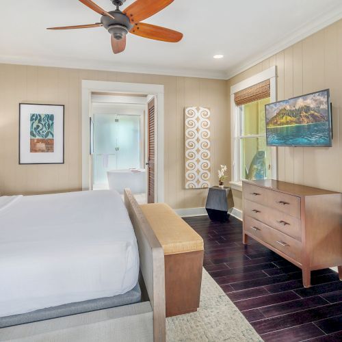 Bright modern bedroom with a bed, dresser, wall TV, windows, and a wooden ceiling fan with warm decor.