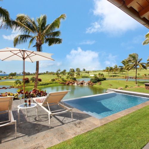 A tropical resort patio with a blue pool, sun loungers, umbrella shade, palm trees, and a pond under a sunny sky.