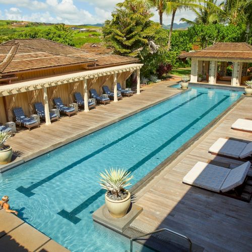 A sunny tropical resort with a long swimming pool, lounge chairs, cabanas, and palm trees surrounding the buildings.