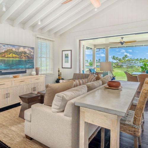 Bright tropical living room with beige sectional sofa, TV on a cabinet, and open patio doors showing lush greenery under ceiling fans.