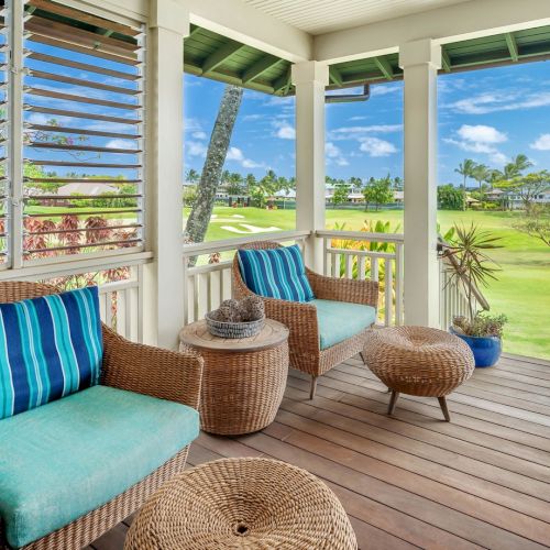 A sunny veranda with wicker lounge chairs and cushions overlooking a lush green golf course and palms under a blue sky.