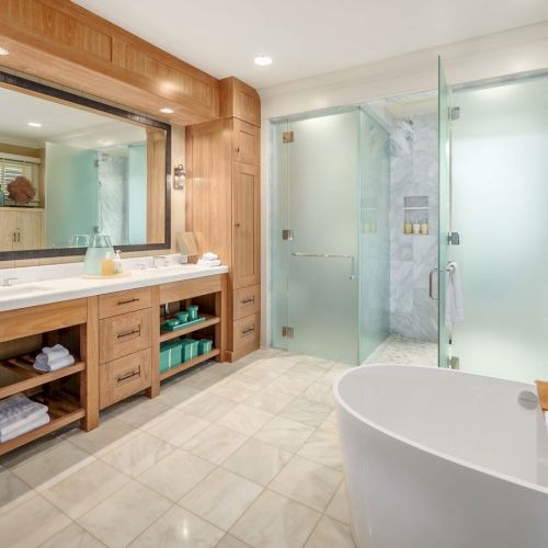 A bright modern bathroom with double vanity, mirror, glass shower, and a white soaking tub with folded towels.