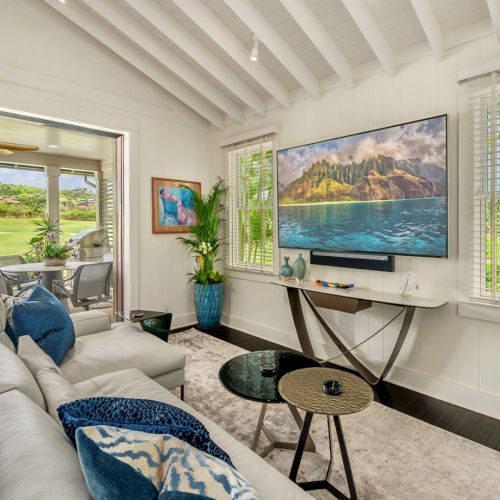 Bright living room with white walls, a sofa, large wall TV, and big windows showing a tropical mountain-and-lake landscape photo.
