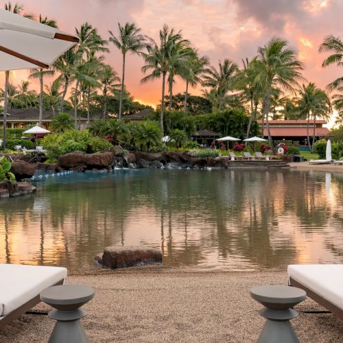 A tropical resort scene with a calm lagoon pool, white loungers, palm trees, and a glowing sunset sky.