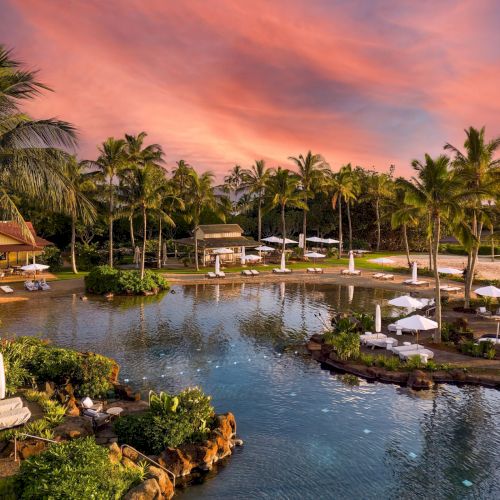 Tropical resort pool at sunset, lined with palm trees, loungers and white umbrellas beside a calm blue waterway.