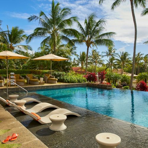 Tropical resort scene: a turquoise swimming pool with sun loungers, umbrellas, and palm trees under a bright sky.