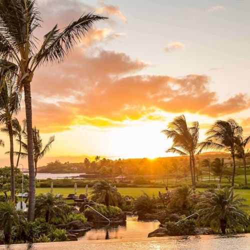 A tropical resort at sunset with tall palm trees, manicured lawns, and a small pond beside the ocean horizon.