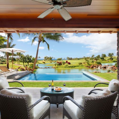A tropical resort patio with two chairs, a small table, a swimming pool, and palm-lined lagoon beyond.