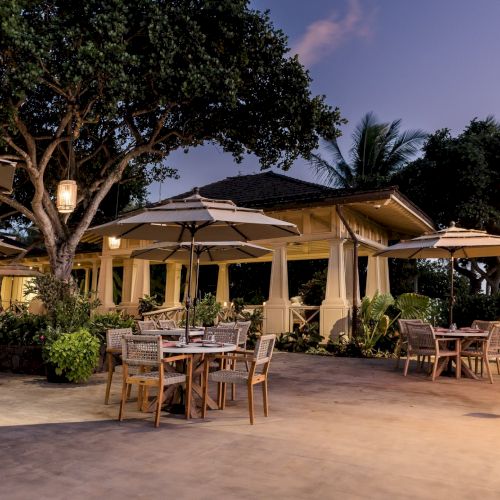A cozy outdoor patio dining area with wicker chairs, tables, large umbrellas, warm lanterns, and palm trees at dusk.