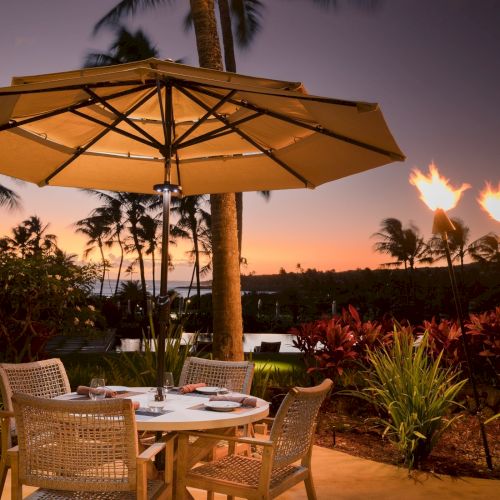 Sunset patio by the ocean with a large orange umbrella shading a table and wicker chairs amid tropical palm trees.