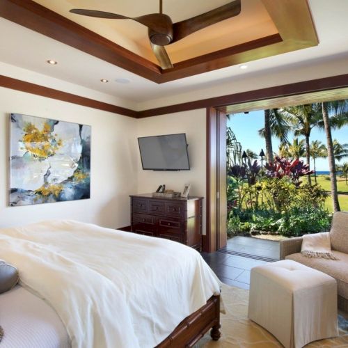 A luxurious bedroom with a ceiling fan, TV, and ocean-view patio doors framed by palm trees outside.