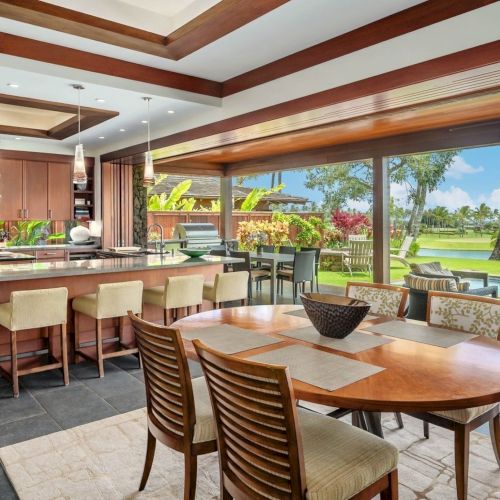 A luxury covered patio with a gourmet kitchen, dining table, bar stools, pool, and lush green lawn in view.