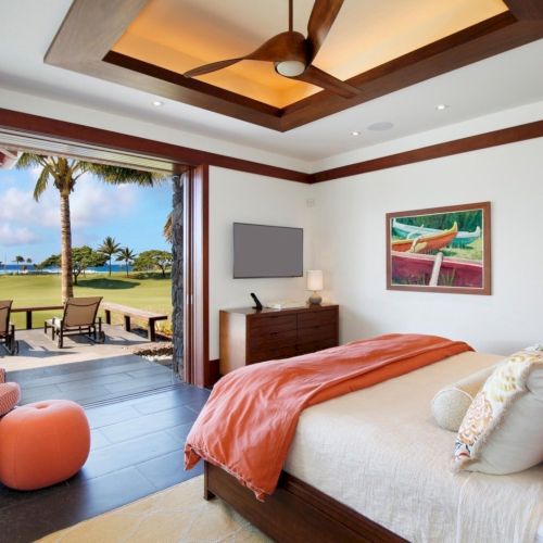 Sunlit bedroom with ocean view, king bed, ceiling fan, TV, and doors opening to a tropical patio with chairs.