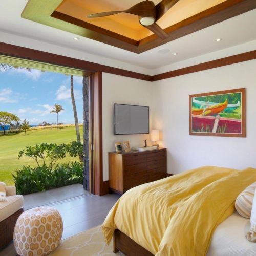 A luxury bedroom with a king bed, ceiling fan, framed art, and large windows showing tropical greenery and ocean views.