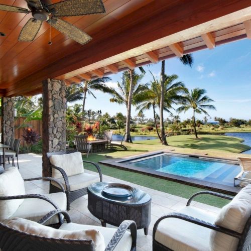 A tropical outdoor patio with a pool, lounge chairs, palm trees, a stone fireplace, and a ceiling fan, overlooking the ocean and greenery.