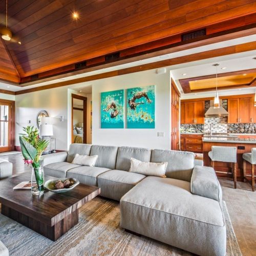 A modern open-plan living room with a gray sectional, a wood-and-stone kitchen bar, pendant lights, and framed wall art.