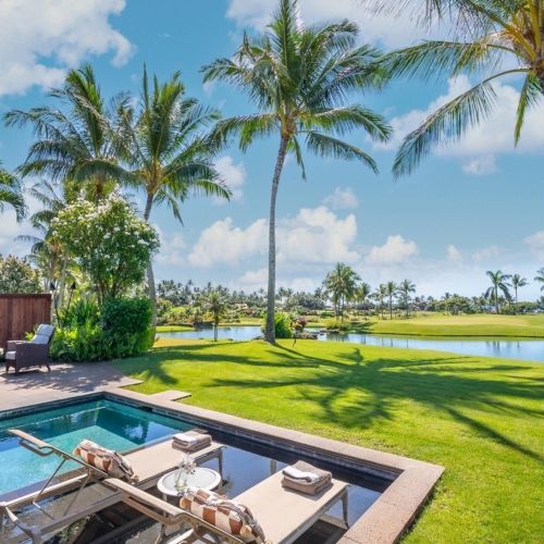 Tropical resort backyard with a swimming pool, sun loungers, lush lawn, and tall palm trees by a calm lake.