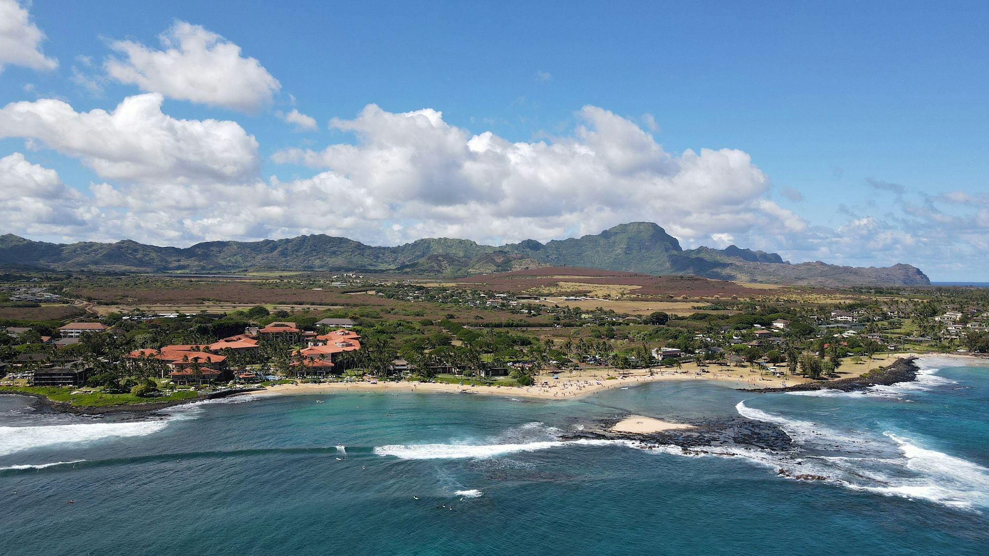 Aerial view of a tropical coastline with a sandy beach, ocean waves, lush town, and mountains under bright clouds.