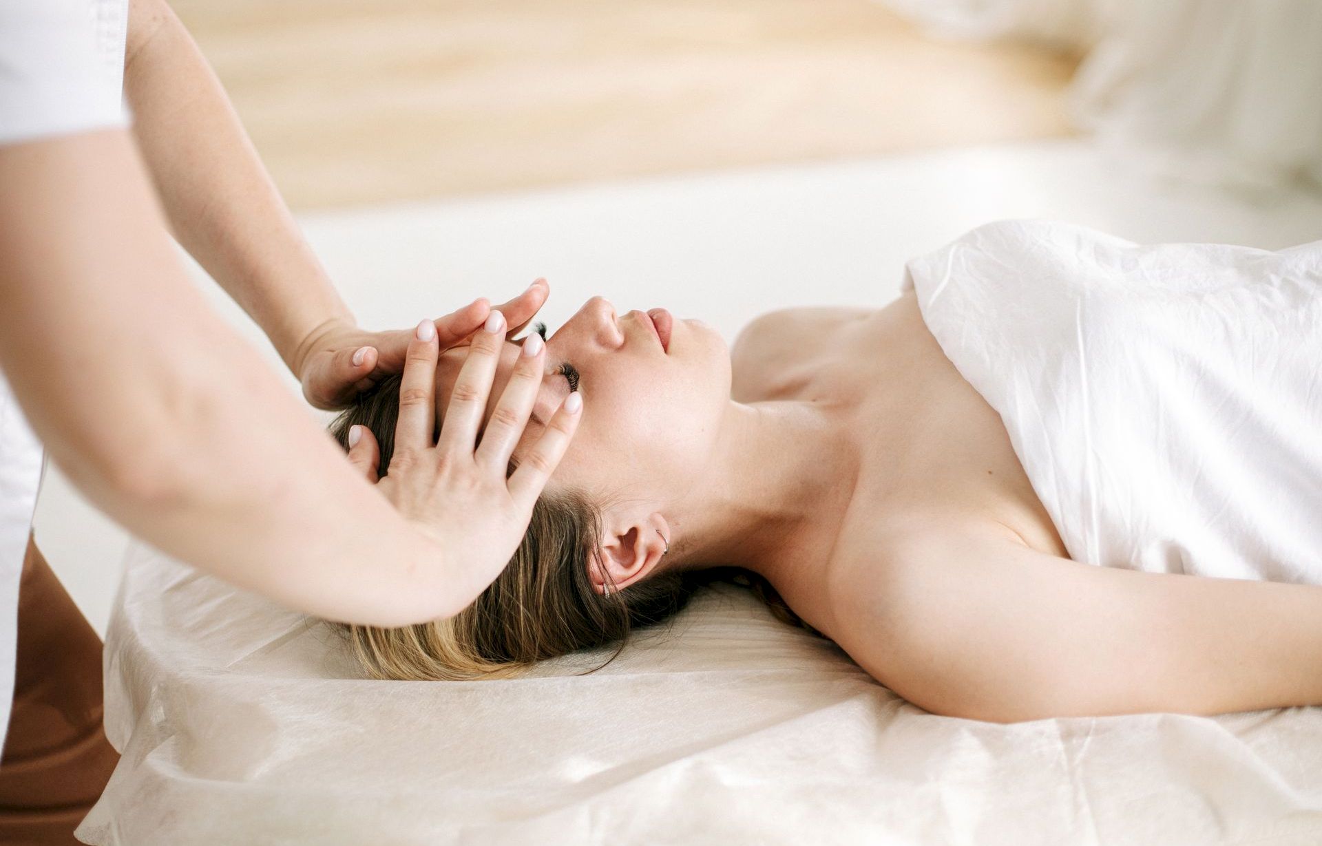 A person giving a gentle facial massage to another while they lie on a bed with soft sheets.