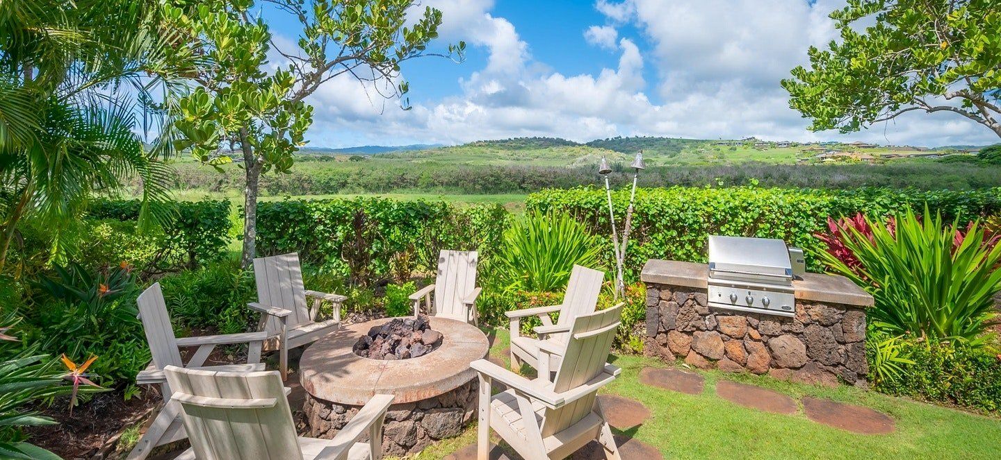 Outdoor patio with white Adirondack chairs around a round firepit, a barbecue grill, and lush green fields under blue sky.
