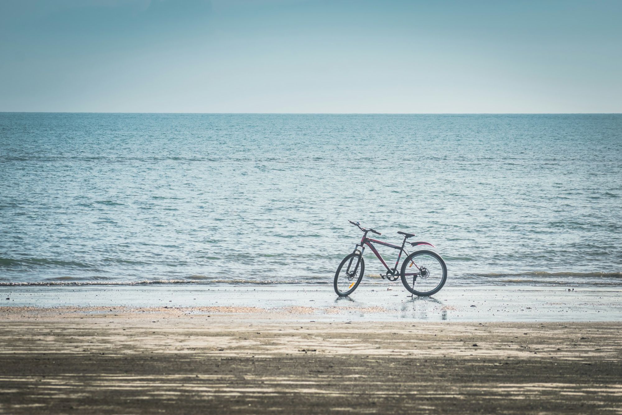 A red bicycle rests on a sandy beach, facing the calm ocean under a clear sky, for an easy seaside ride.